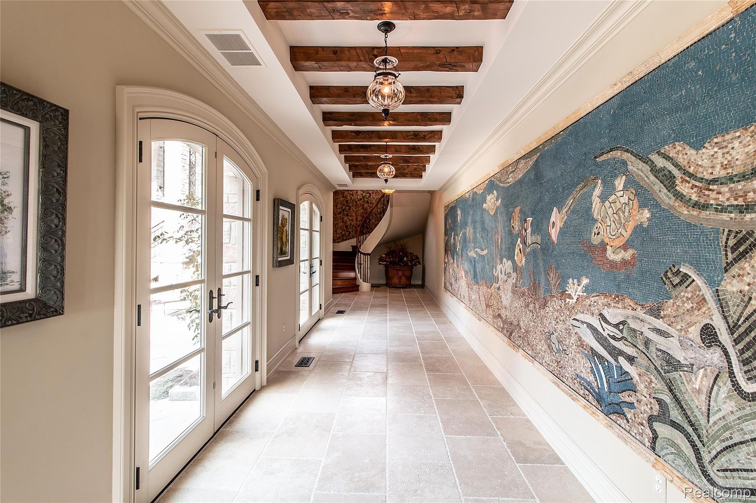 This interior shot showcases a grand hallway featuring a mosaic wall, wooden ceiling beams with pendant lighting, and arched French doors leading to the exterior. The hallway is spacious with light-colored stone flooring, leading to a curved staircase in the background. The overall impression is luxurious and artistic.