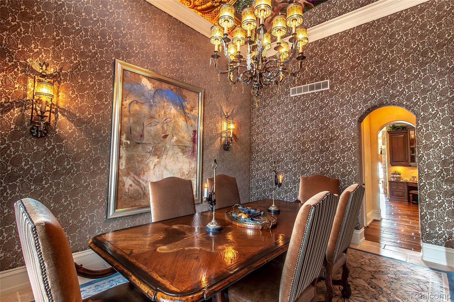 This is an interior shot of a formal dining room. The room features a large wooden dining table with upholstered chairs, an ornate chandelier, and patterned wallpaper. A large painting hangs on the wall, and there are sconces providing additional lighting. The room exudes a sense of luxury and traditional elegance.