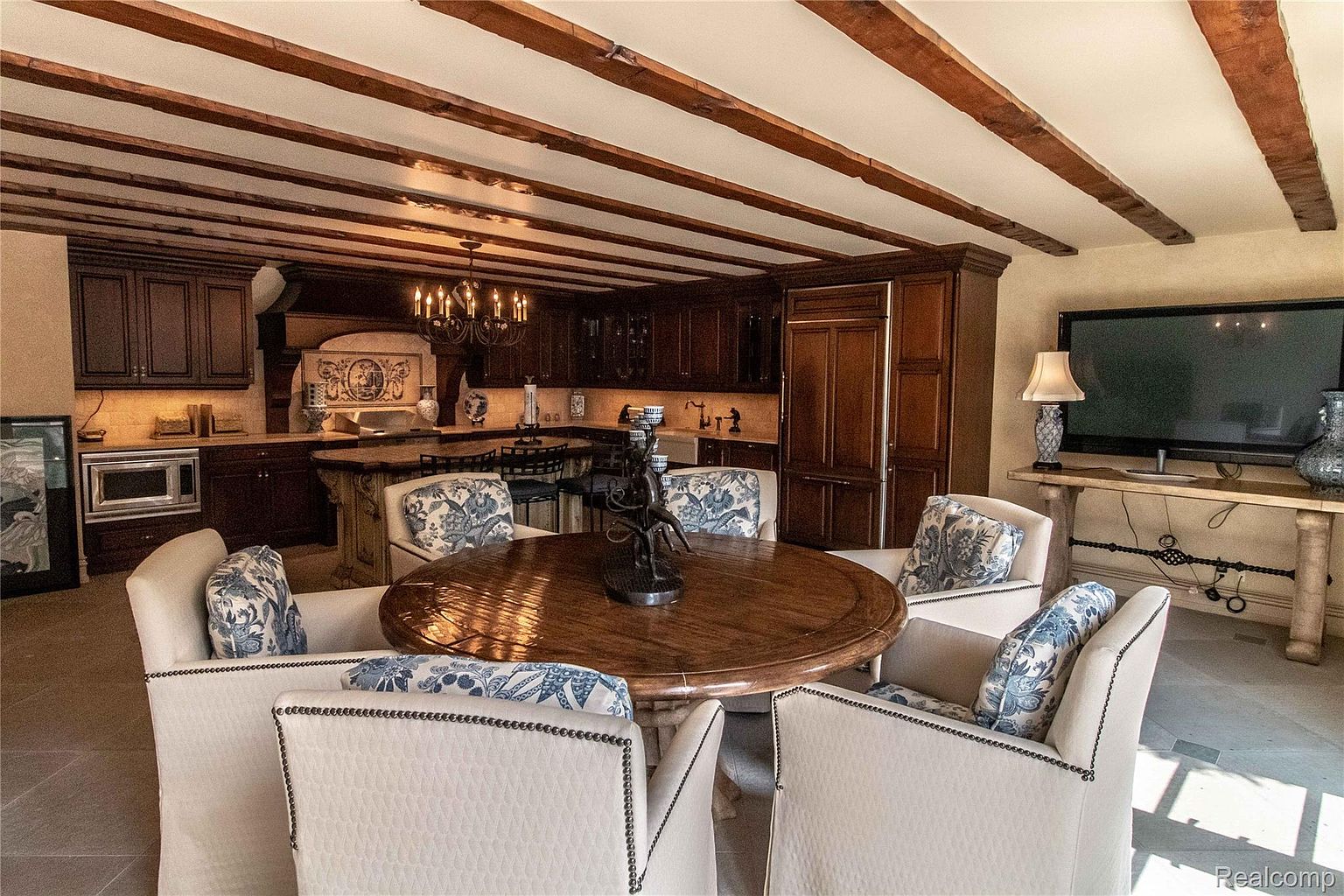 This interior shot showcases a dining area adjacent to a kitchen, featuring a large round wooden table surrounded by six upholstered chairs with nailhead trim. The room is characterized by dark wood cabinetry in the kitchen area, exposed wooden beams on the ceiling, and a large television on a console table, creating a warm and inviting atmosphere.