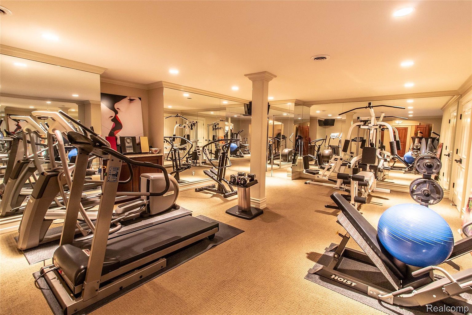 This is a well-equipped home gym featuring various exercise machines, including treadmills, weight machines, and free weights. The room is spacious with mirrored walls, enhancing the sense of size and providing visual feedback during workouts. The neutral carpet and lighting create a comfortable and functional workout environment.