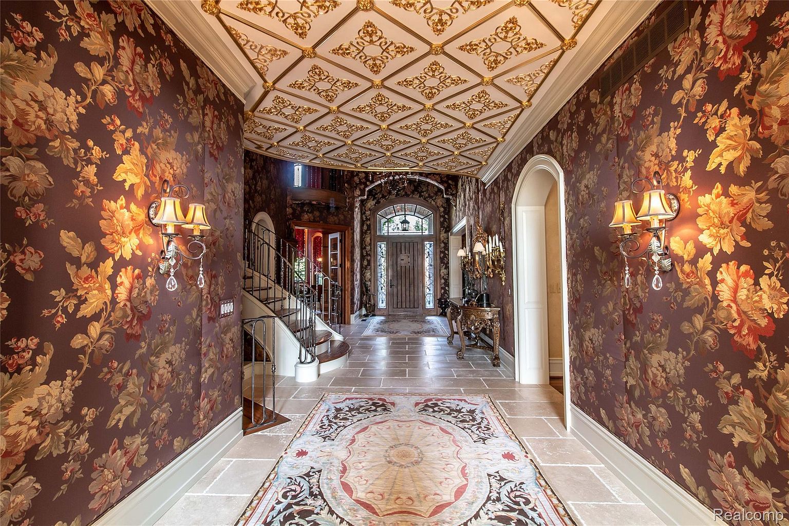 This is an interior shot of a grand hallway featuring ornate floral wallpaper, a coffered ceiling with gold detailing, and a patterned rug on a stone floor. A staircase is visible to the left, and an arched doorway leads further into the home, creating a luxurious and inviting entrance. The lighting from the sconces adds warmth and highlights the intricate details of the design.