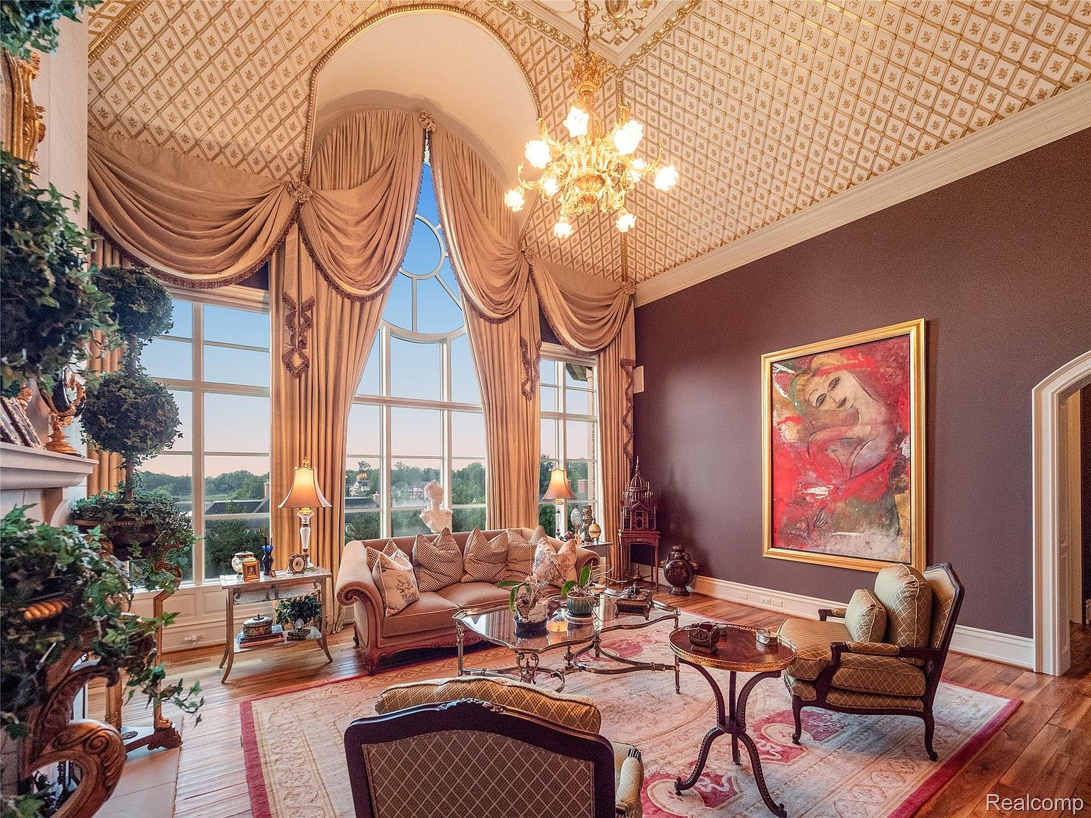 This is an opulent living room featuring a soaring arched window with elaborate drapery, a decorative ceiling with gold accents, and a large painting on a dark brown wall. The room is furnished with a plush sofa, ornate armchairs, and glass-topped tables, all arranged on an area rug with a classic pattern. The perspective is from a low angle, emphasizing the height and grandeur of the space.