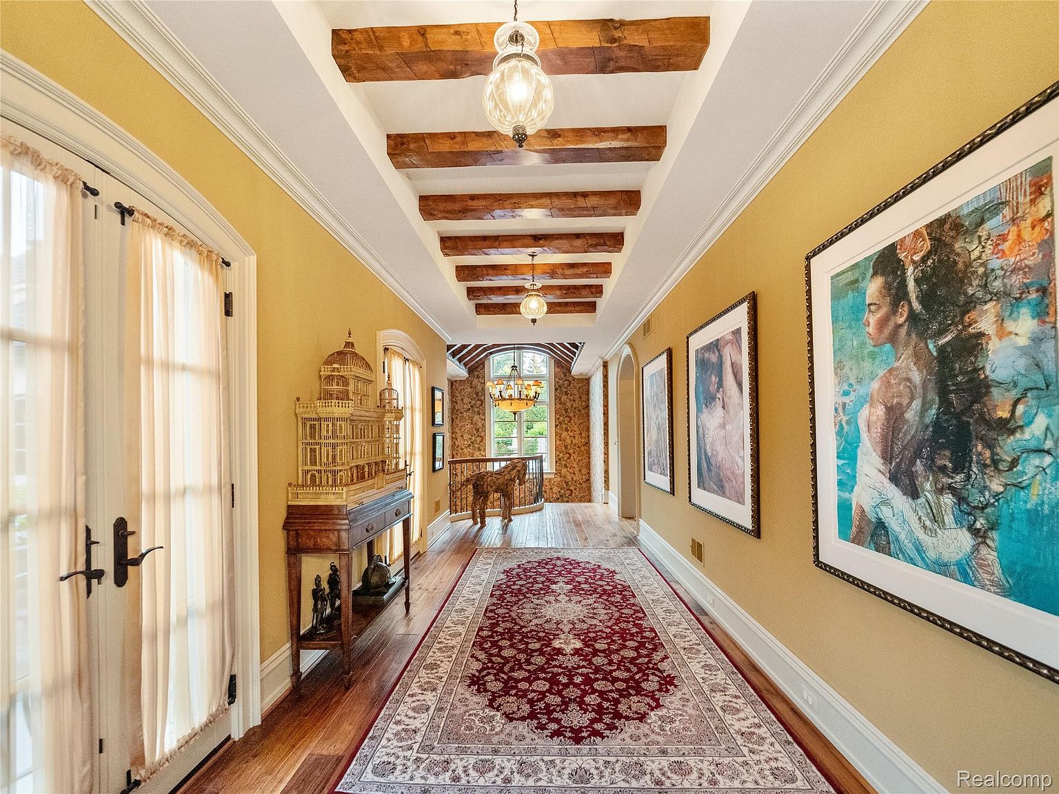 This interior shot showcases a grand hallway with rich architectural details. The hallway features wooden beams on the ceiling, warm yellow walls adorned with framed artwork, and a patterned rug that adds a touch of elegance. A decorative birdhouse-like structure sits on a table to the left, and a glimpse of a staircase with a horse statue can be seen at the end of the hallway, creating a sense of depth and luxury.