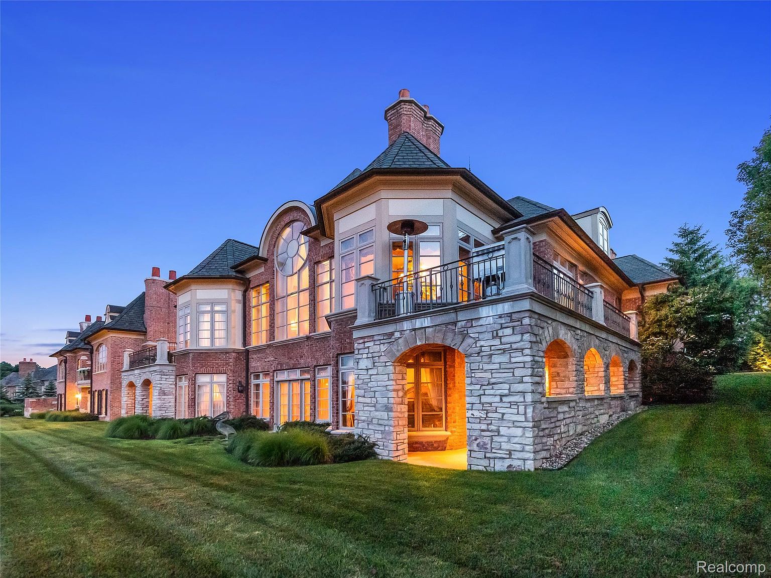 This is a stunning front view of a large, luxurious home at dusk. The house features a combination of brick and stone facade, multiple windows with warm interior lighting, and a prominent balcony. The manicured lawn and landscaping add to the property's curb appeal, creating an inviting and impressive estate.