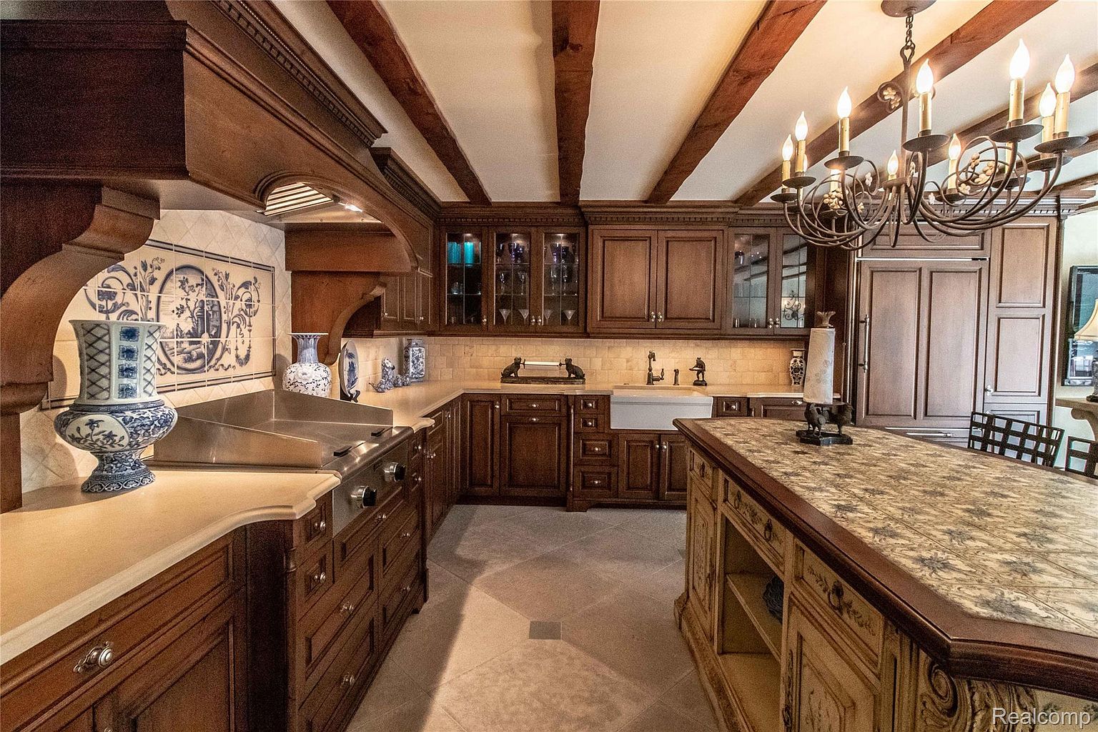 This is a well-appointed kitchen featuring dark wood cabinetry, a large center island with a decorative countertop, and a farmhouse sink. The kitchen also boasts a professional-grade range with a custom hood and decorative tile backsplash. The ceiling has exposed wooden beams and a decorative chandelier, adding to the room's luxurious and traditional style.