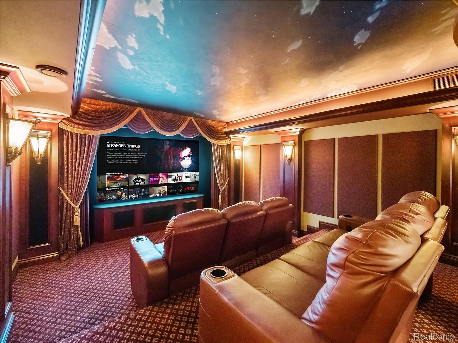 This is an interior shot of a home theater. The room features tiered seating with leather recliners, a large screen displaying "Stranger Things", and custom lighting that mimics a starry night sky on the ceiling. The walls are paneled with dark wood and fabric, creating a cozy and immersive viewing experience.