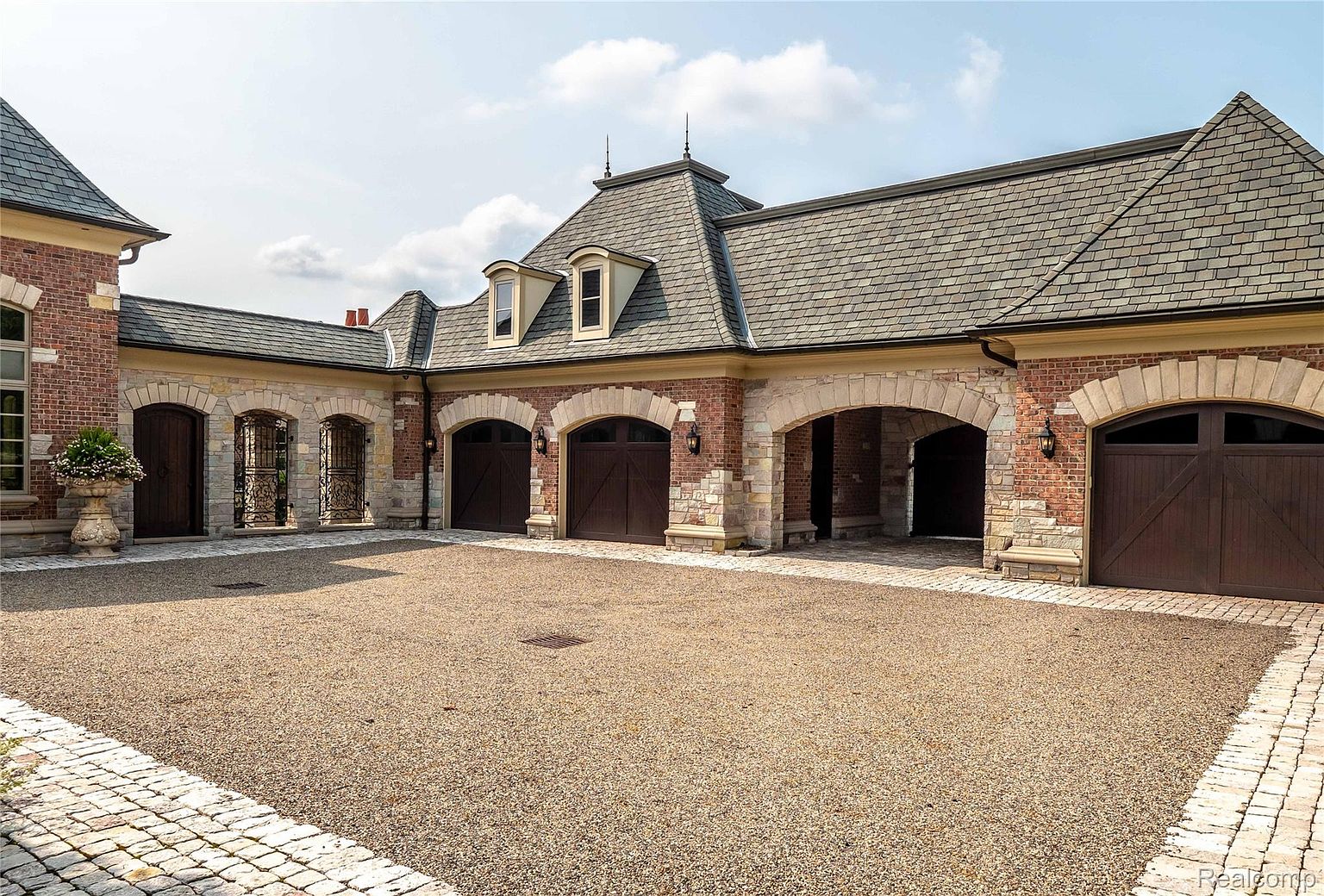 This image showcases a luxurious garage exterior, constructed with red brick and stone accents, featuring multiple arched entrances and dark wooden garage doors. The driveway is covered in gravel, bordered by cobblestone, adding to the estate's sophisticated and timeless appeal. The roof is made of grey shingles and the overall design exudes high-end craftsmanship and architectural detail.