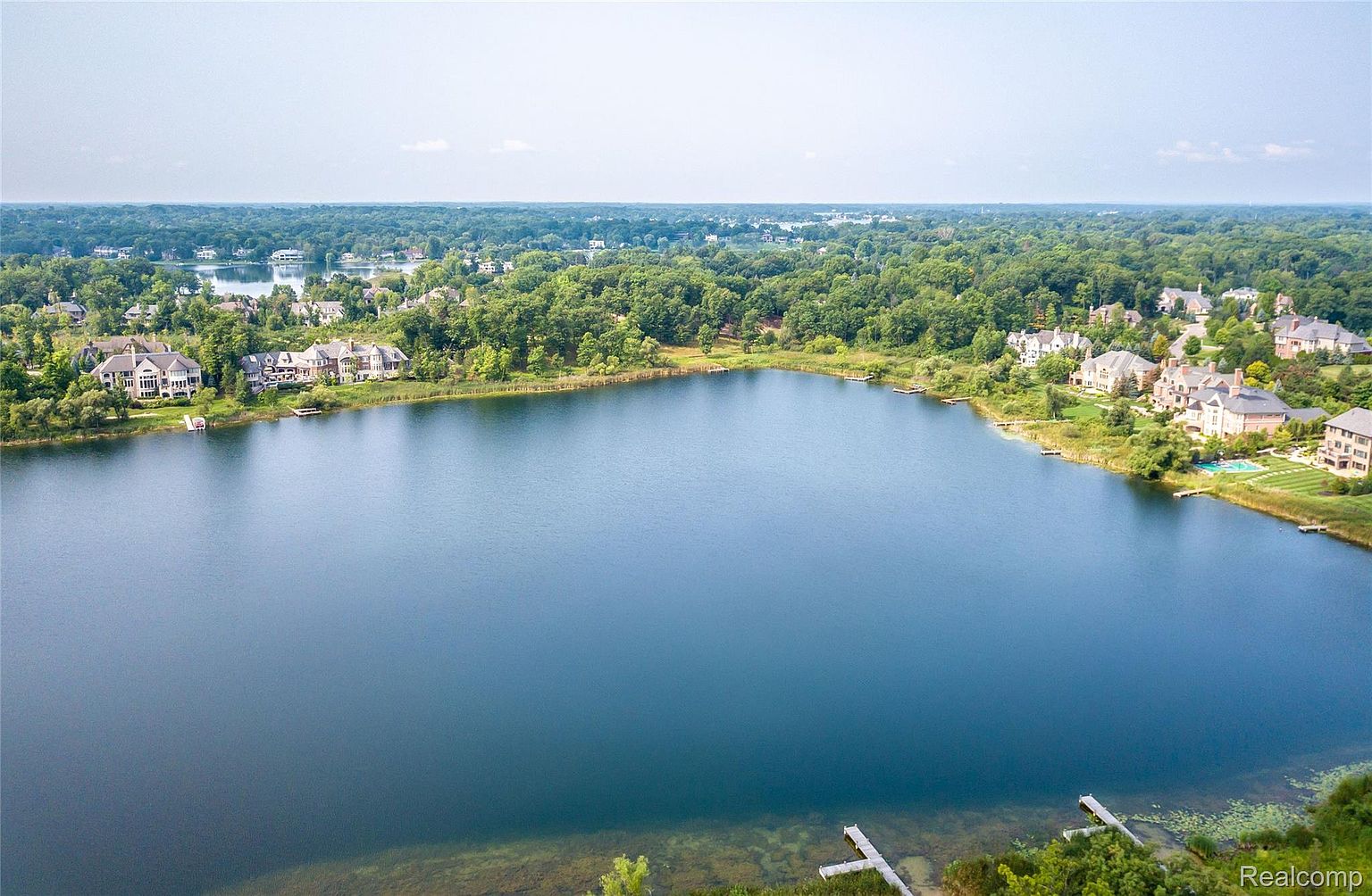 This aerial view showcases a serene lake surrounded by lush greenery and upscale residential properties. The homes feature varied architectural styles, some with pools and docks, adding to the luxurious lakeside living experience. The overall impression is one of tranquility and high-end real estate.