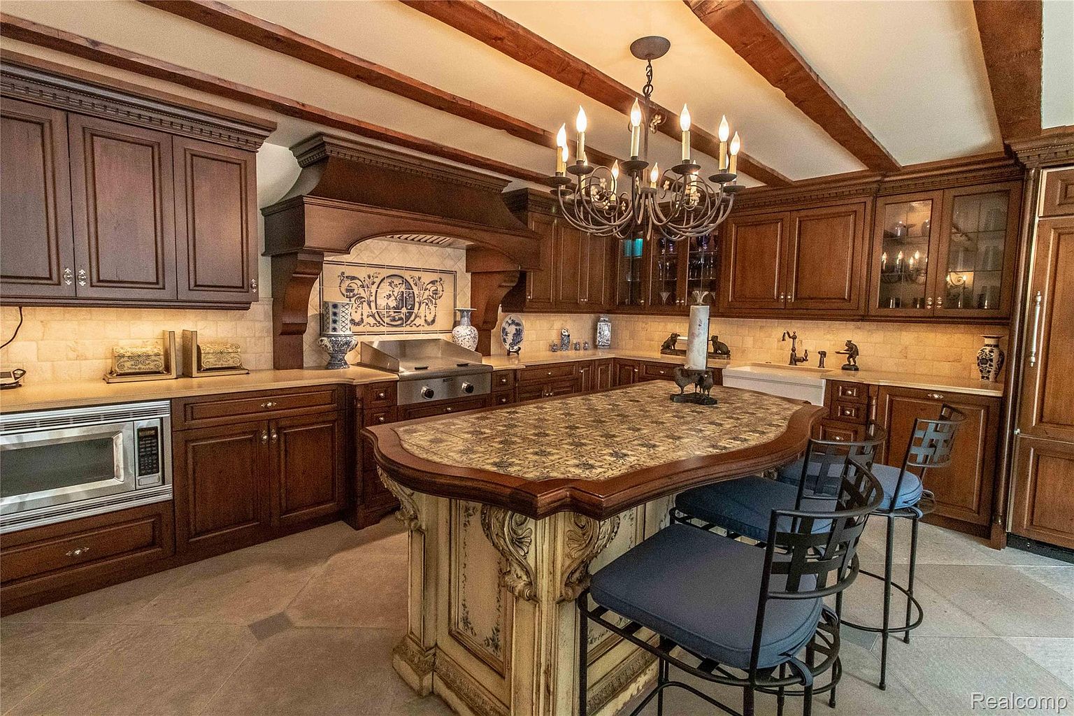 This is a well-appointed kitchen featuring dark wood cabinetry, a decorative tile backsplash above the range, and a large island with seating. The island has a unique patterned countertop and ornate detailing on the base. A chandelier hangs above the island, adding a touch of elegance to the space.