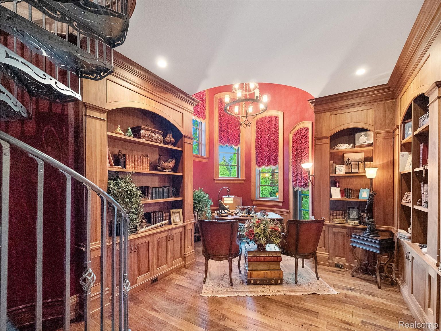 This is an interior shot of a luxurious home office or study. The room features custom wood built-in bookshelves, a central desk with chairs, and a spiral staircase. The walls are painted a rich red, and the room is illuminated by a chandelier, creating a warm and inviting atmosphere.