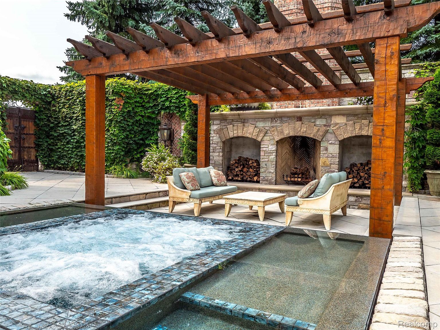 This image showcases a luxurious outdoor spa area featuring a hot tub and a smaller pool, both with tiled edges. A wooden pergola provides shade over a seating area with comfortable chairs and a small table, set against a stone fireplace with wood storage niches. Lush greenery surrounds the space, creating a private and serene atmosphere.