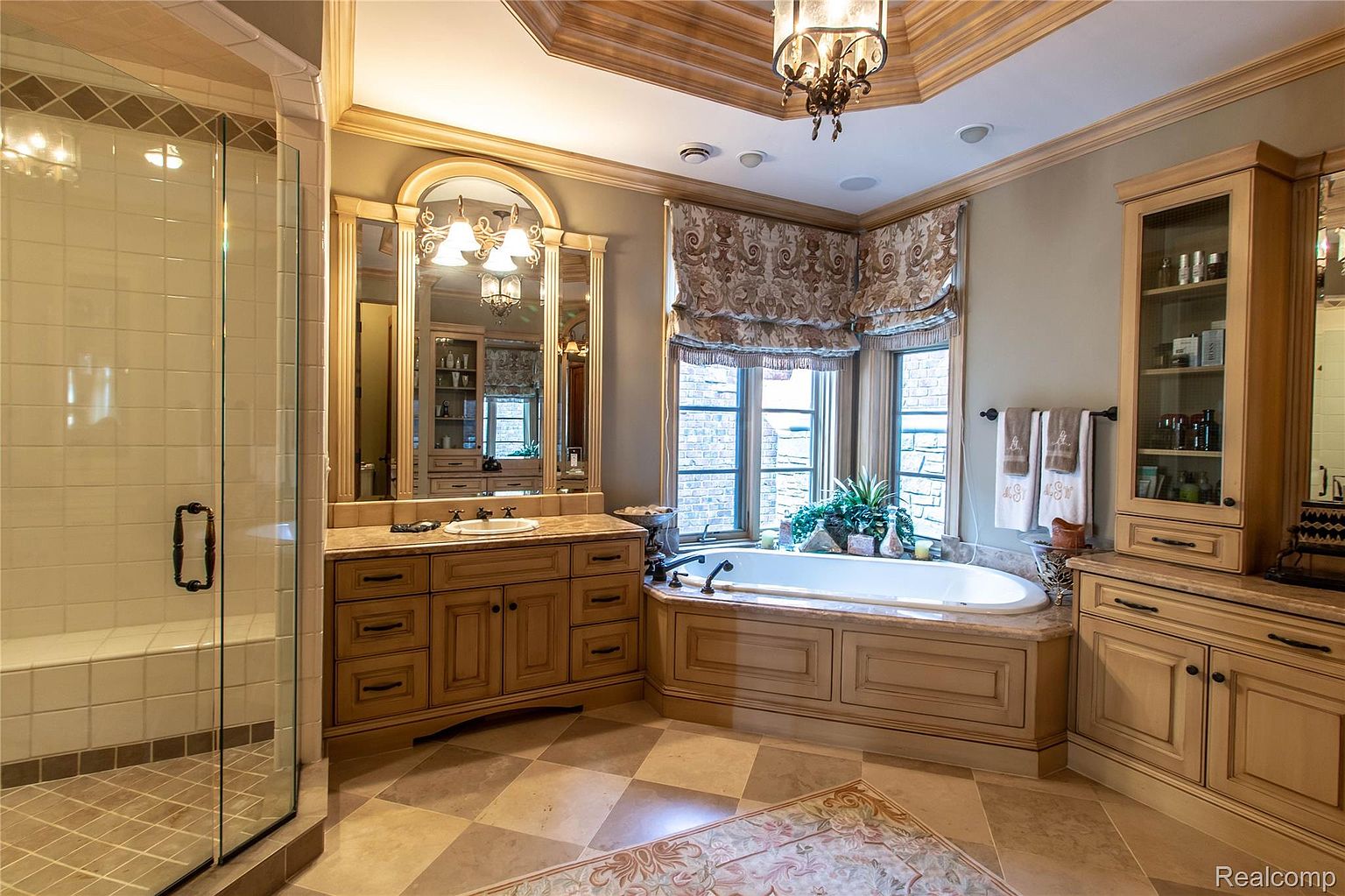 This is a luxurious primary bathroom featuring a large vanity with a marble countertop and ornate mirror, a soaking tub surrounded by windows with custom blinds, and a glass-enclosed shower. The room is decorated in neutral tones with elegant fixtures and finishes, creating a spa-like atmosphere. The flooring is a patterned tile, and there is a built-in cabinet for storage.