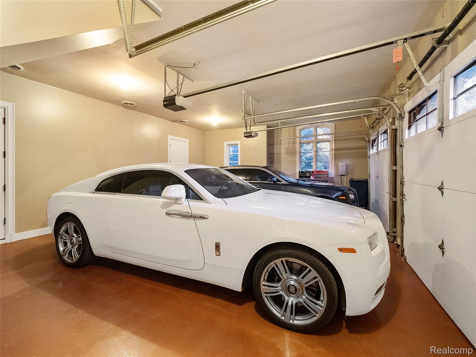 This is an interior shot of a well-lit garage featuring two luxury cars, one white and one dark. The garage has a clean, organized appearance with a painted floor and a modern garage door with windows. The walls are painted in a neutral tone, and the overall impression is one of luxury and functionality.