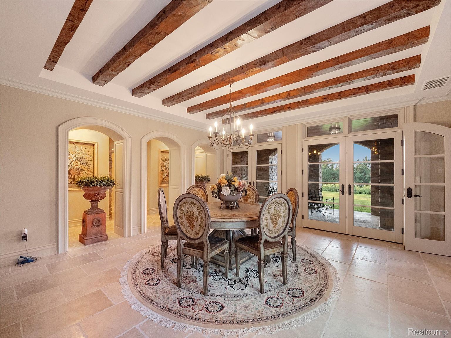This is an elegant dining room featuring a round wooden table surrounded by upholstered chairs on a circular rug. The room has exposed wooden beams on the ceiling, a chandelier, and arched doorways leading to other rooms. French doors offer a view of the outdoor space, creating a bright and inviting atmosphere.