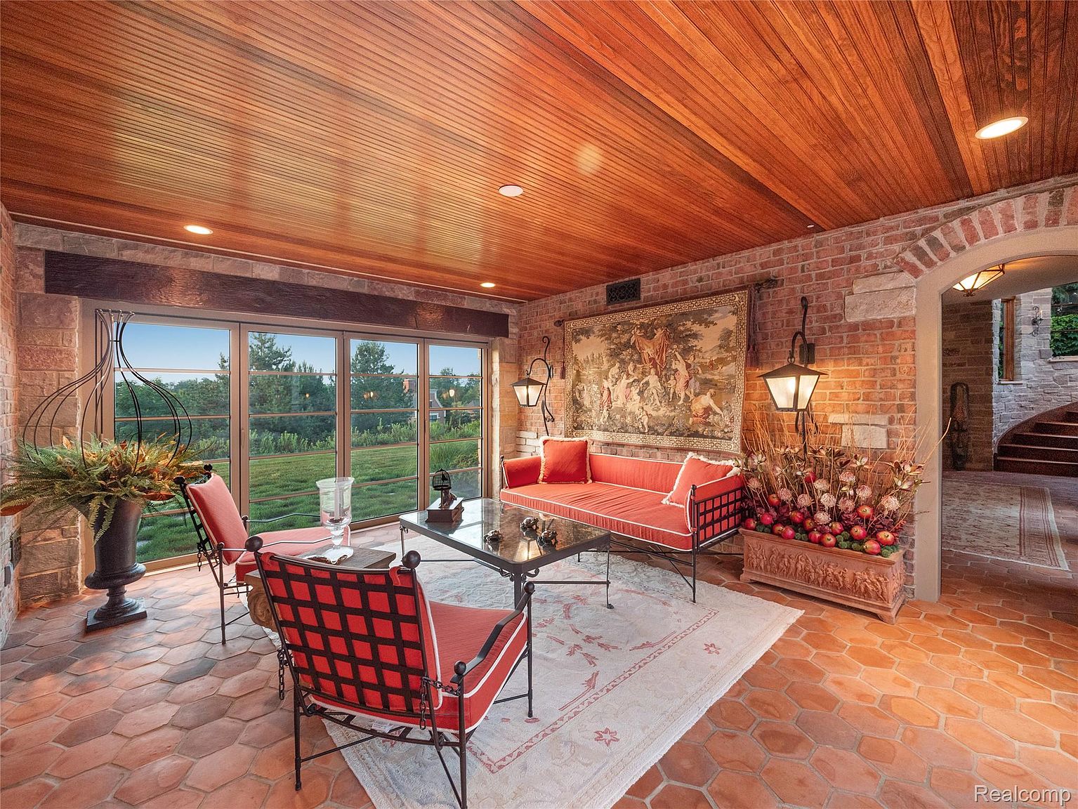 This sunroom features a warm and inviting atmosphere with a wood-paneled ceiling and brick walls. The room is furnished with a red sofa and chairs, a glass-topped coffee table, and a decorative rug. Large windows offer a view of the outdoor landscape, and the terracotta tile flooring adds to the room's rustic charm.
