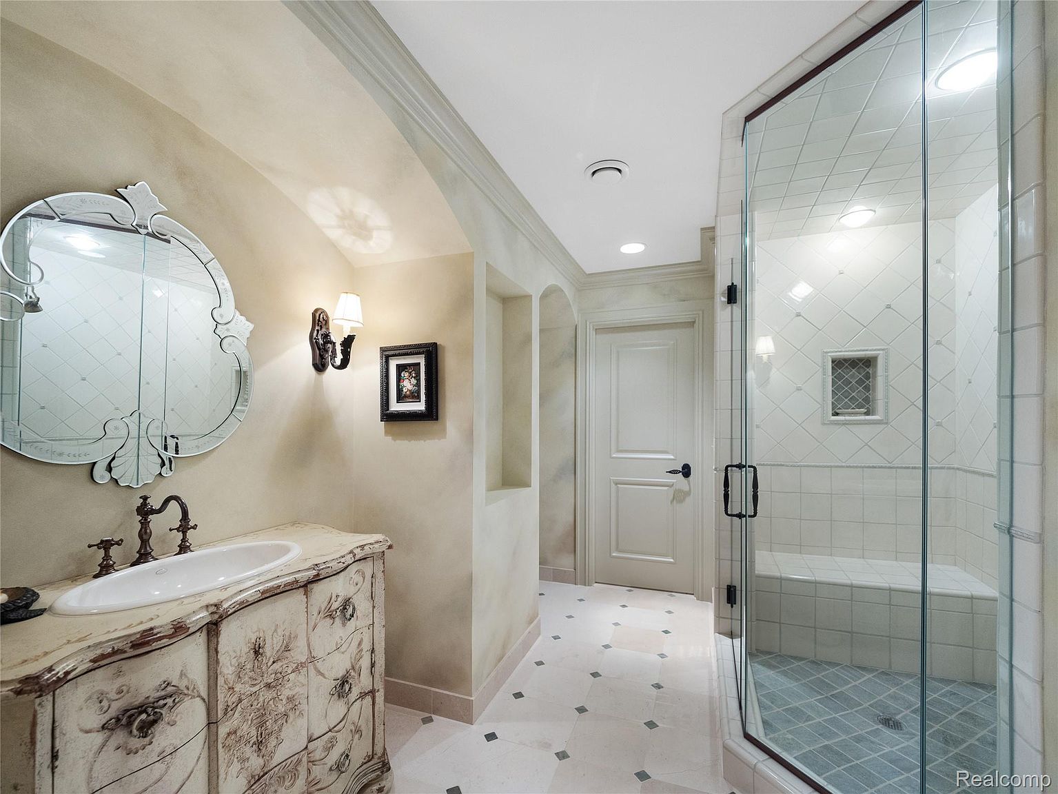 This primary bathroom features a decorative vanity with an oval sink and ornate mirror. The shower is enclosed with glass and has a built-in bench and tiled walls. The flooring is tiled with a decorative pattern, and the overall style is elegant and traditional.