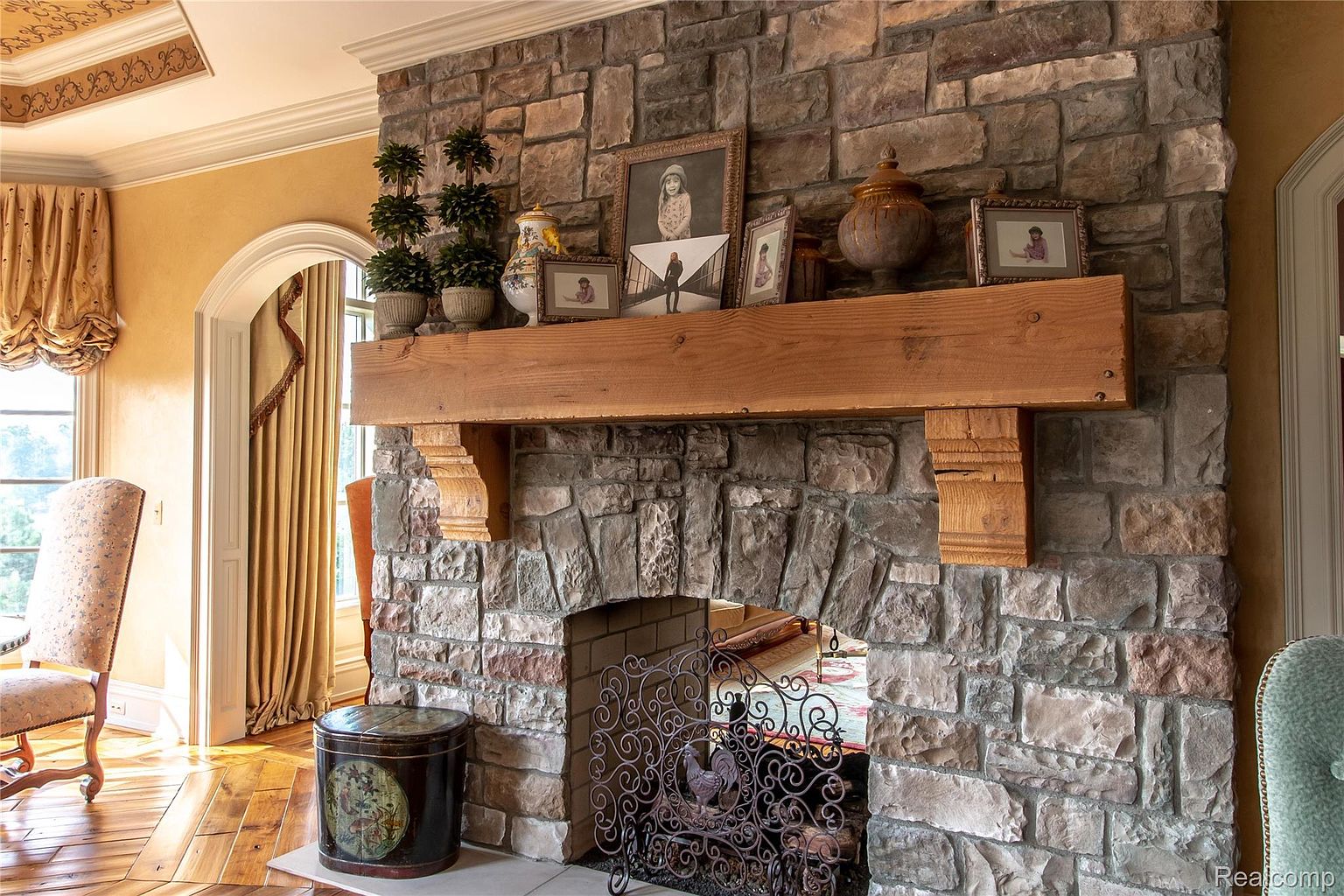 This interior shot showcases a grand stone fireplace with a wooden mantel, adorned with decorative items and framed pictures. The fireplace features an arched opening with a decorative metal screen. The room exudes a traditional and luxurious ambiance, enhanced by the hardwood flooring and elegant furnishings visible in the periphery.
