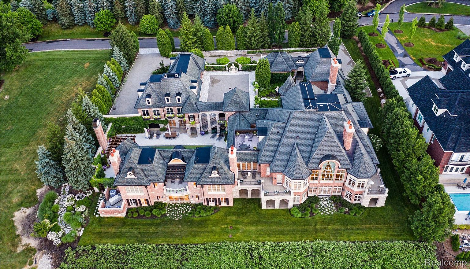This is an aerial view of a large, luxurious estate. The property features a sprawling mansion with a complex roofline, multiple chimneys, and a mix of brick and stone facades. The meticulously landscaped grounds include manicured lawns, mature trees, and formal gardens, creating an impression of grandeur and privacy.