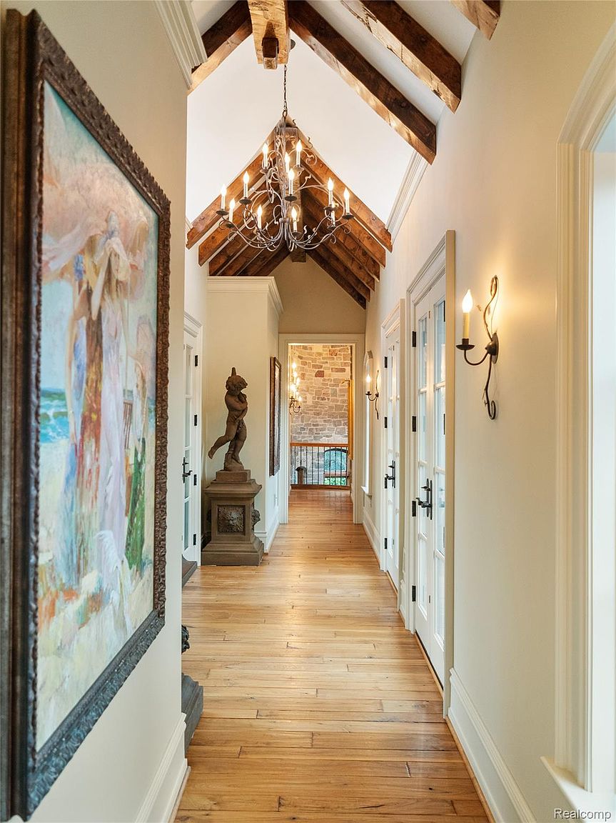 This is an interior shot of a hallway featuring hardwood floors, a vaulted ceiling with exposed wooden beams, and a decorative chandelier. A large framed painting hangs on the left wall, and a statue stands on a pedestal further down the hall. The hallway is well-lit with sconces and natural light, creating a warm and inviting atmosphere.