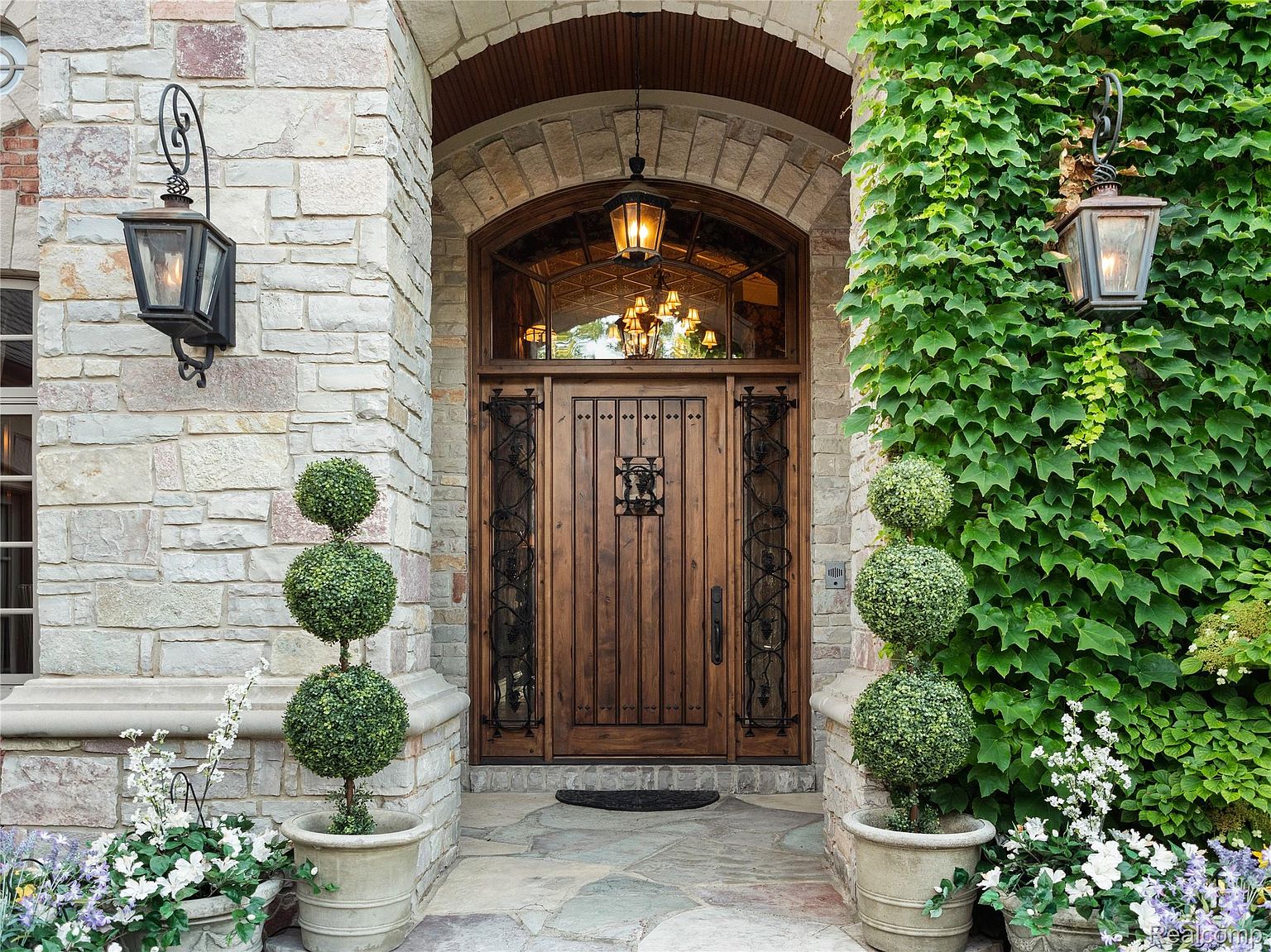 This is a front entryway shot of a luxurious home. The focal point is a grand wooden door with decorative ironwork, framed by a stone archway and lush greenery. Elegant lanterns flank the entrance, adding to the home's curb appeal and creating a welcoming atmosphere.