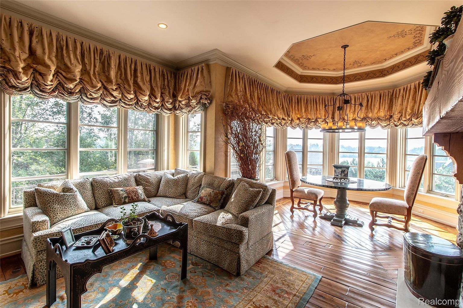 This sunroom features a large bay window with decorative valances, providing ample natural light and views of the surrounding landscape. A patterned sectional sofa and a round table with chairs create a cozy seating area. The hardwood flooring and decorative ceiling add to the room's elegant and inviting atmosphere.