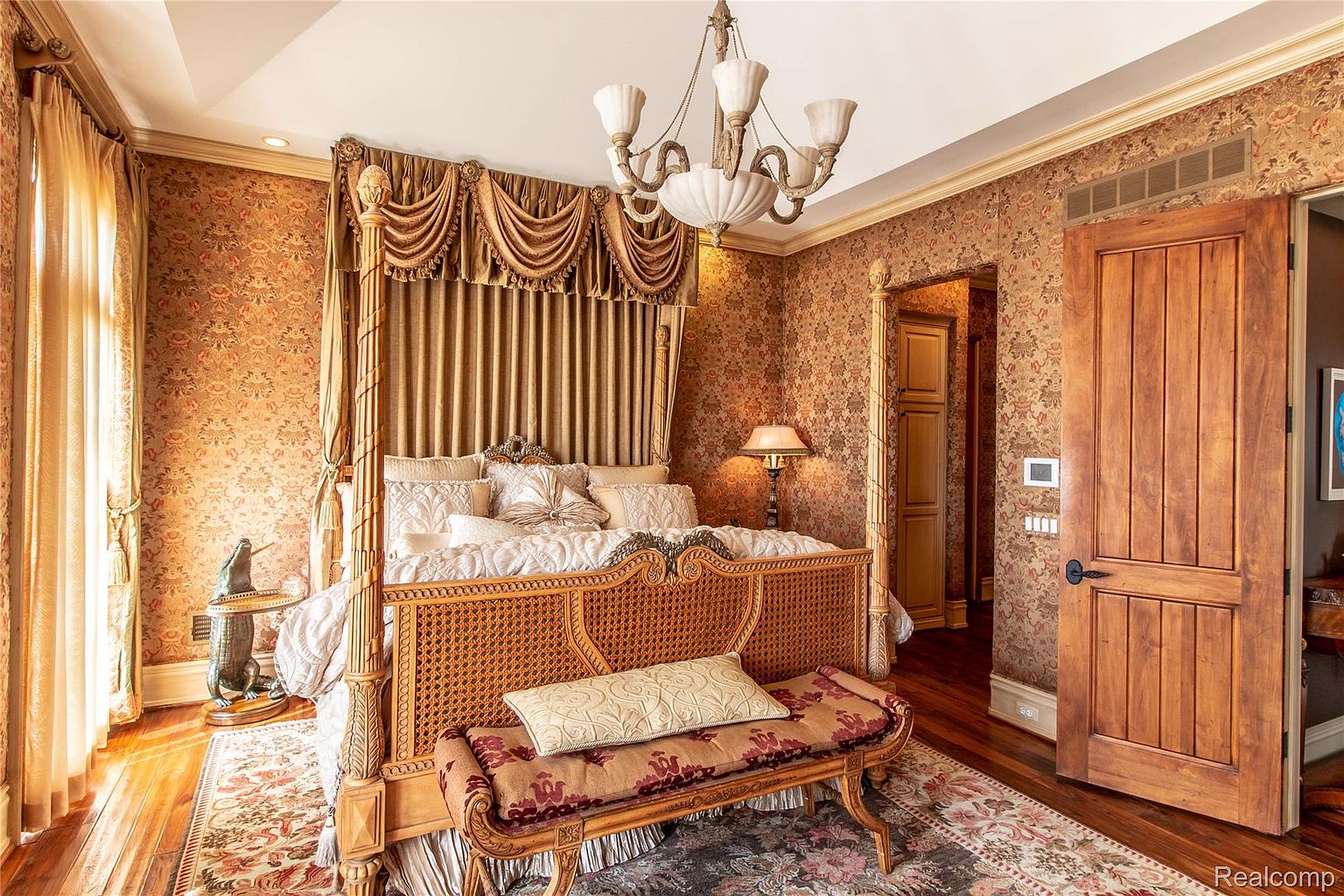 This is a luxurious primary bedroom featuring a four-poster bed with ornate detailing and gold drapery. The room is decorated with patterned wallpaper and a complementary rug, creating a rich and elegant atmosphere. A chandelier hangs from the ceiling, adding to the room's opulence.