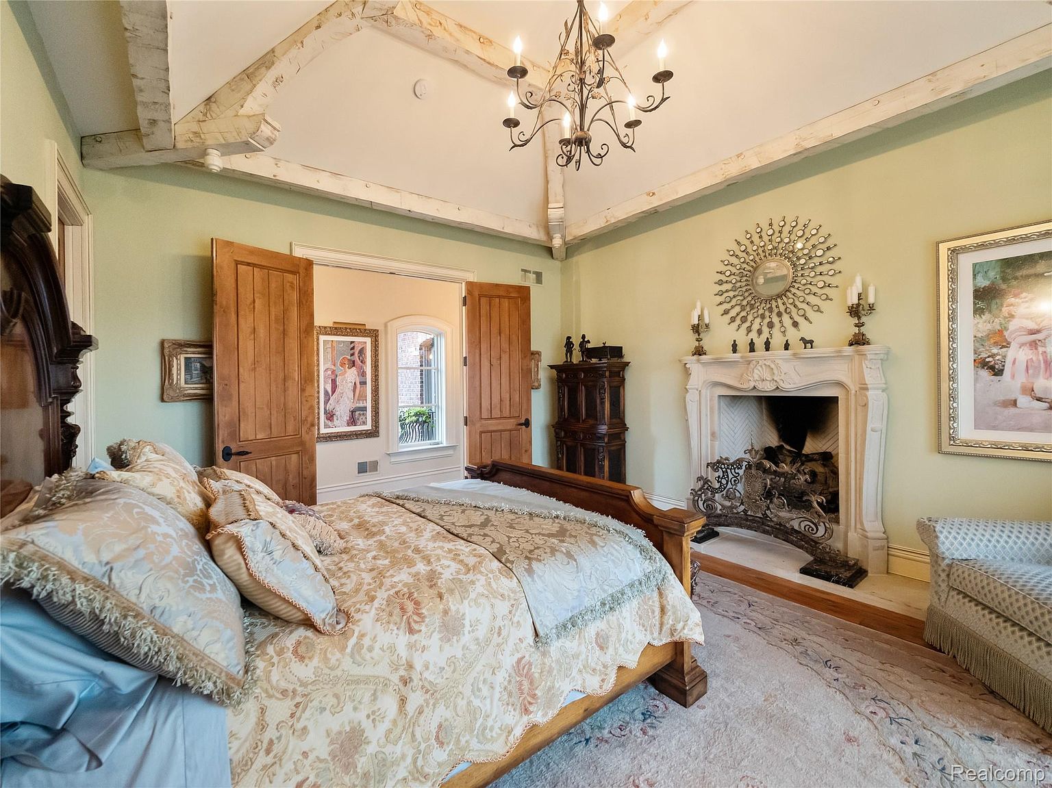 This is a primary bedroom featuring a large bed with decorative pillows and a patterned comforter. The room has a fireplace with an ornate mantel, a chandelier, and wooden beams on the ceiling. The walls are painted a soft green, creating a warm and inviting atmosphere.
