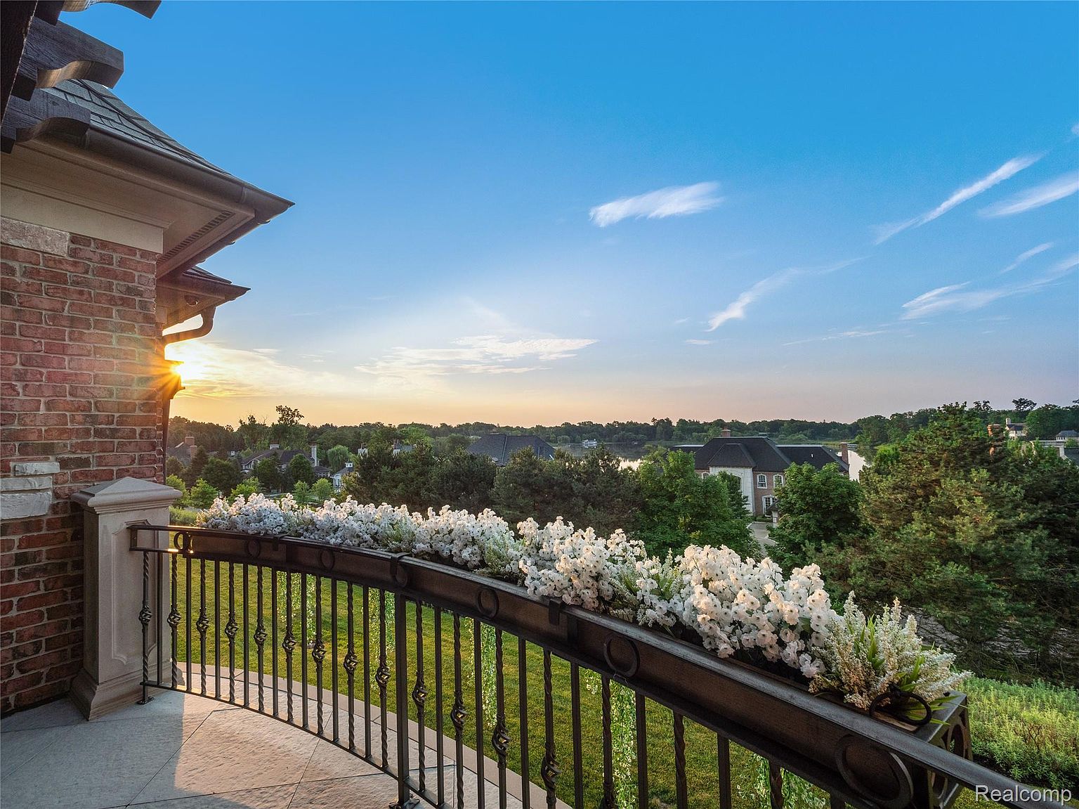 This image showcases a luxurious balcony with an ornate wrought-iron railing adorned with white flowers. The balcony offers a scenic view of a lush green landscape with trees and a glimpse of a lake, all under a beautiful sky at sunset. The architectural details of the building, including the brickwork and roofline, add to the overall elegance and charm of the property.