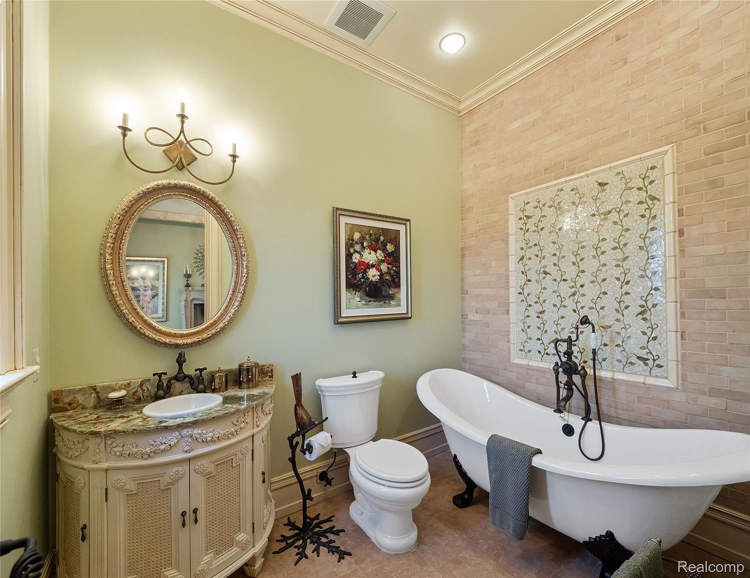 This is a primary bathroom featuring a clawfoot bathtub, an ornate vanity with a marble countertop and a round mirror above it. The walls are painted a light green color, with one wall featuring brick-like tiles and a decorative mosaic panel. The overall style is luxurious and classic.