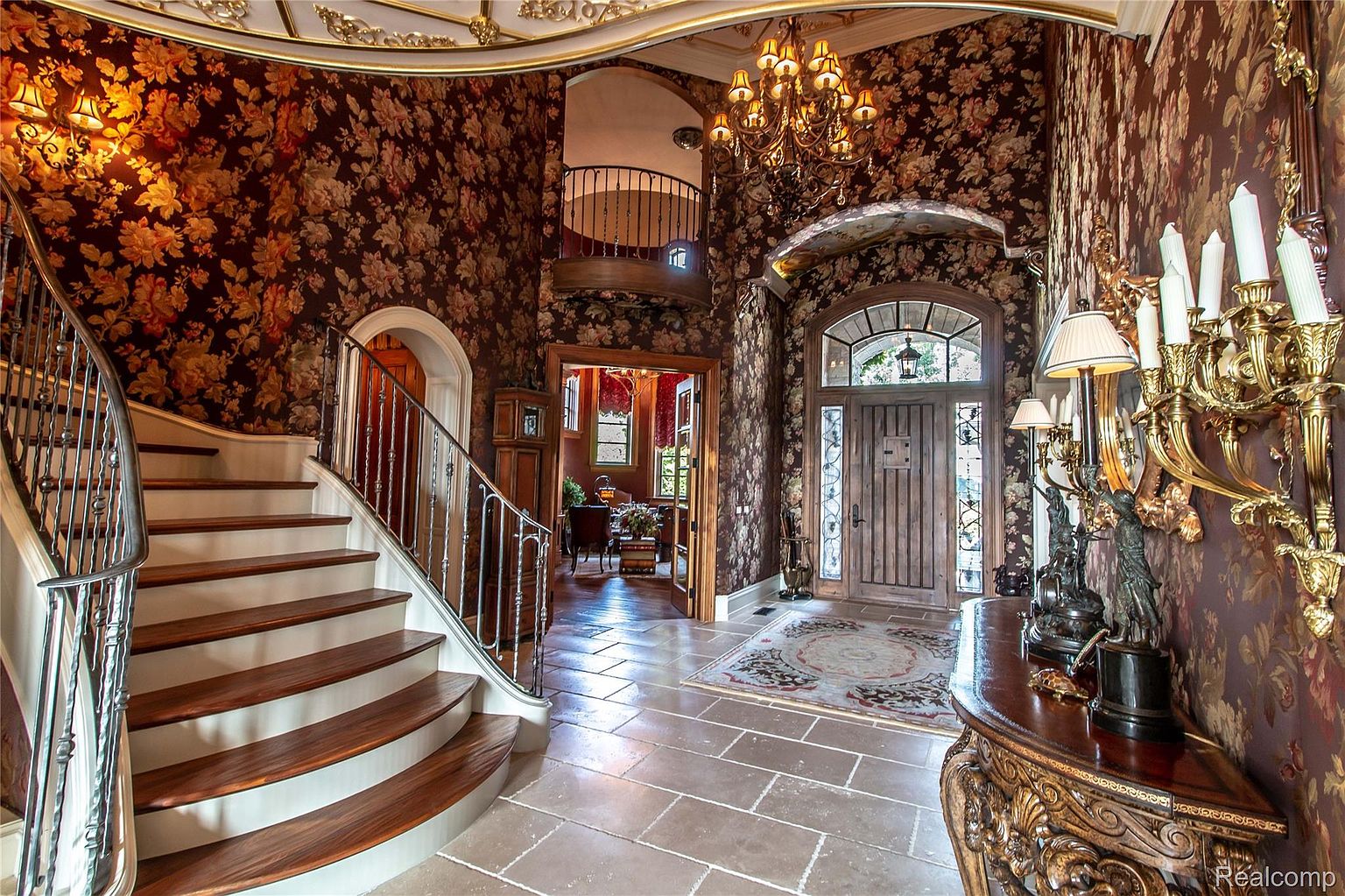 This grand foyer features a curved staircase with wooden treads and white risers, complemented by an ornate metal railing. The walls are adorned with a rich floral wallpaper, and a large chandelier hangs from the ceiling, adding to the luxurious ambiance. The flooring is composed of large stone tiles, and a doorway leads to another room, creating a sense of depth and flow.