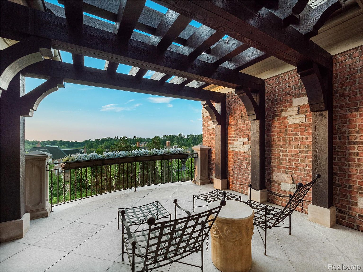 This image showcases a luxurious outdoor balcony or patio area. The space features a dark wood pergola, brick columns with stone accents, and elegant metal furniture. The balcony overlooks a lush green landscape, creating a serene and inviting atmosphere, perfect for relaxation and outdoor entertaining.
