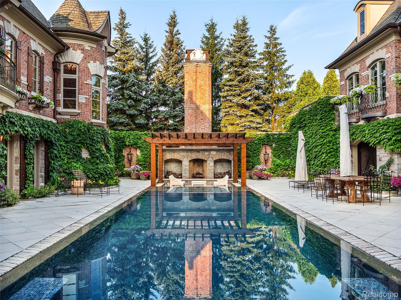 This image showcases a luxurious outdoor pool area, featuring a long, rectangular pool with a clear reflection of a brick chimney and pergola. The pool is surrounded by a stone patio, lush greenery, and ivy-covered walls, creating a private and elegant retreat. The architectural style of the adjacent buildings is traditional with brick facades and detailed windows.