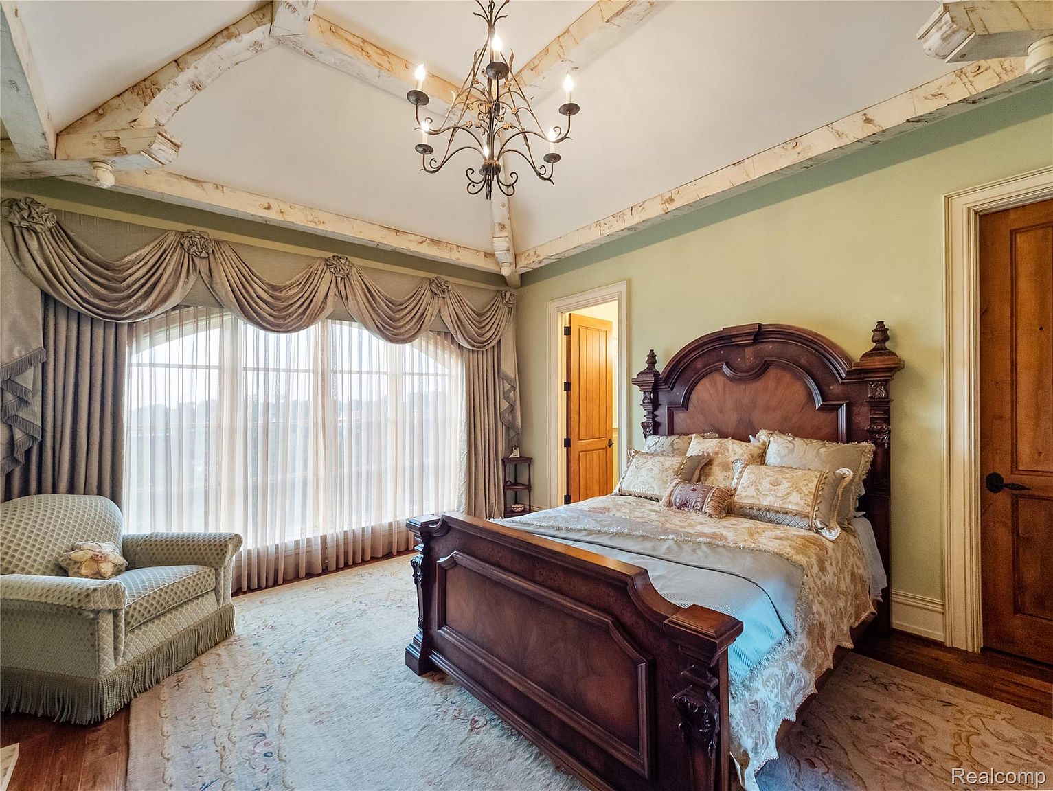 This is a primary bedroom featuring a large, ornate wooden bed with decorative pillows and a patterned bedspread. The room has a light green wall color, a large window with elegant drapery, and a unique ceiling with exposed wooden beams and a chandelier. The overall impression is luxurious and traditional.