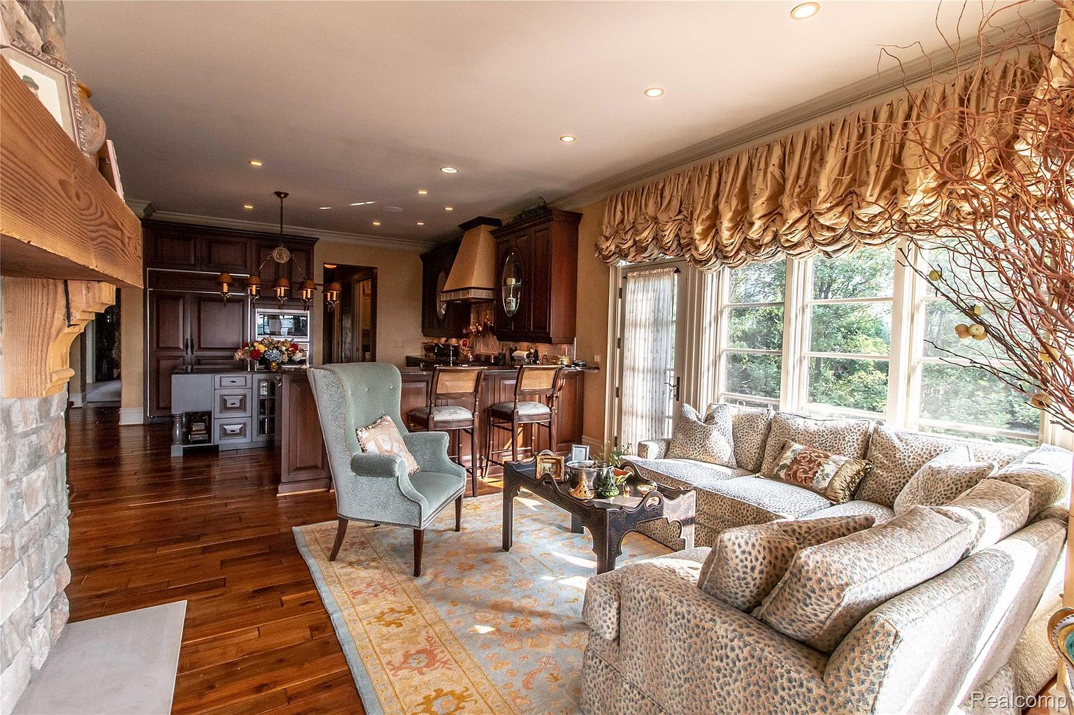 This is an interior shot of a living room that seamlessly blends into a kitchen area. The living room features a patterned sofa, a light blue armchair, and a decorative rug, creating a cozy and inviting atmosphere. The kitchen area includes dark wood cabinetry and bar seating, suggesting a modern and functional space for cooking and entertaining.