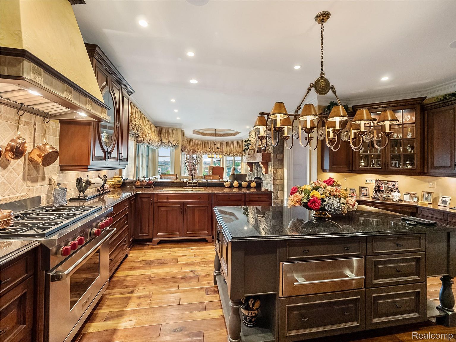 This is a spacious, luxurious kitchen featuring dark wood cabinetry, granite countertops, and stainless steel appliances. A large island with a black countertop and a decorative chandelier above it serves as a focal point. The hardwood floors and warm lighting create an inviting and elegant atmosphere.