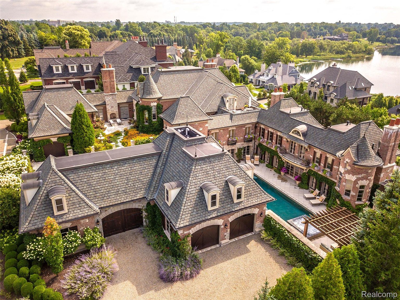 This aerial shot showcases a luxurious estate with multiple interconnected buildings, featuring a cohesive architectural style with brick facades and gray shingle roofs. A swimming pool and meticulously landscaped gardens add to the property's appeal, while a nearby lake provides a scenic backdrop. The overall impression is one of grandeur and sophisticated living.