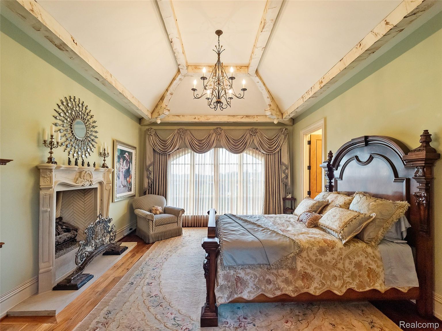 This is a luxurious primary bedroom featuring a grand wooden bed with ornate detailing and plush bedding. The room includes a fireplace with an elaborate mantel and a decorative mirror above. A large window with elegant drapery provides natural light, and the overall style is opulent and traditional.