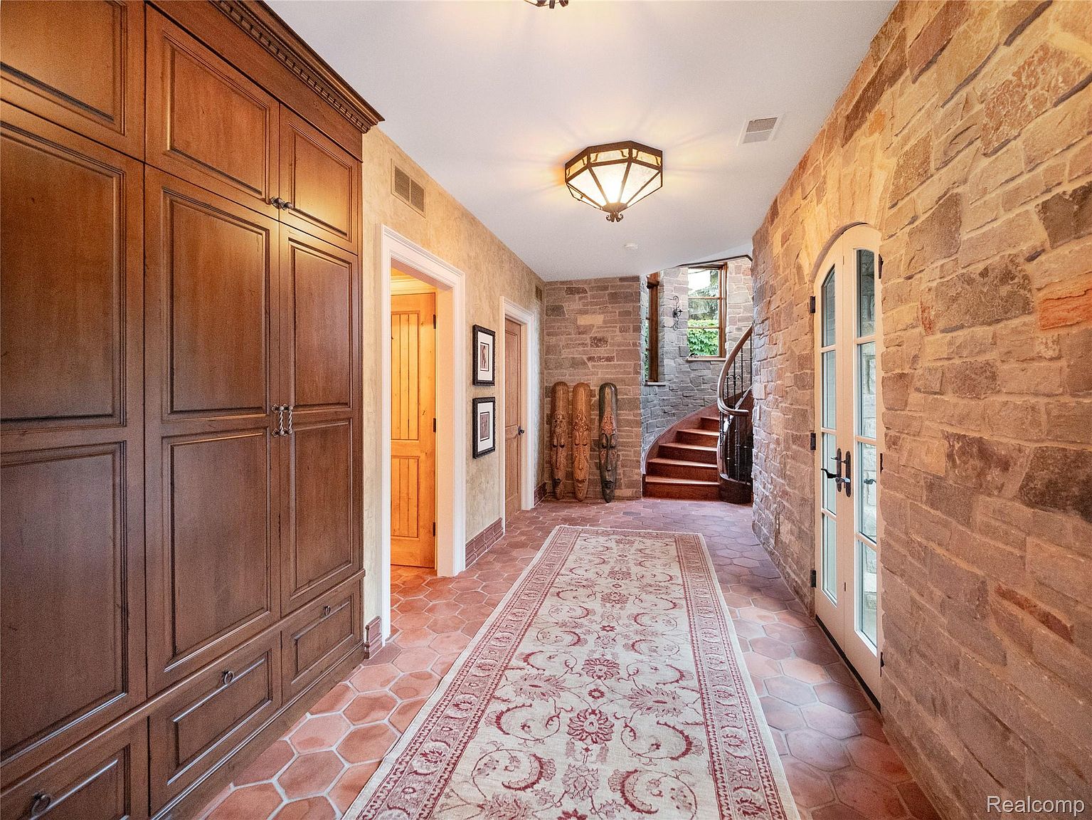 This interior shot showcases a hallway with unique architectural details. The hallway features a combination of stone and textured walls, complemented by a warm-toned tile floor and a patterned rug. A wooden staircase with a curved banister adds a touch of elegance, while built-in wooden cabinets provide ample storage space.