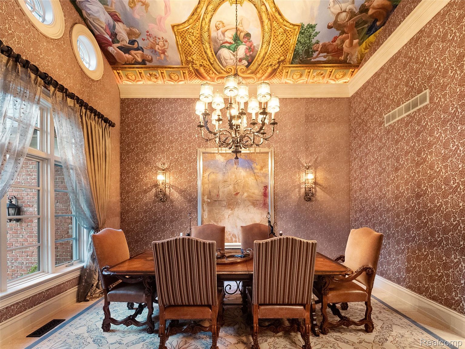 This is an interior shot of a formal dining room. The room features a large wooden dining table surrounded by upholstered chairs, a decorative chandelier, and an ornate ceiling mural. The walls are covered in patterned wallpaper, and the room is lit by sconces and natural light from a window, creating a luxurious and elegant atmosphere.
