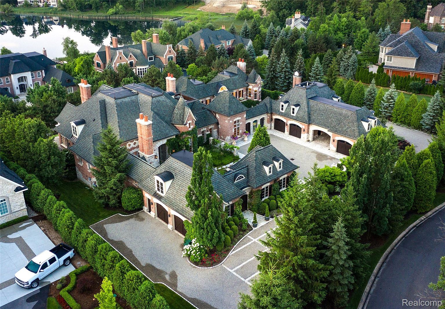 This is an aerial view of a large, luxurious estate with multiple interconnected buildings, including a main house and several garage structures. The property features meticulously landscaped grounds with mature trees, manicured hedges, and a circular driveway. The architectural style is traditional with brick and stone accents, and the overall impression is one of grandeur and privacy.