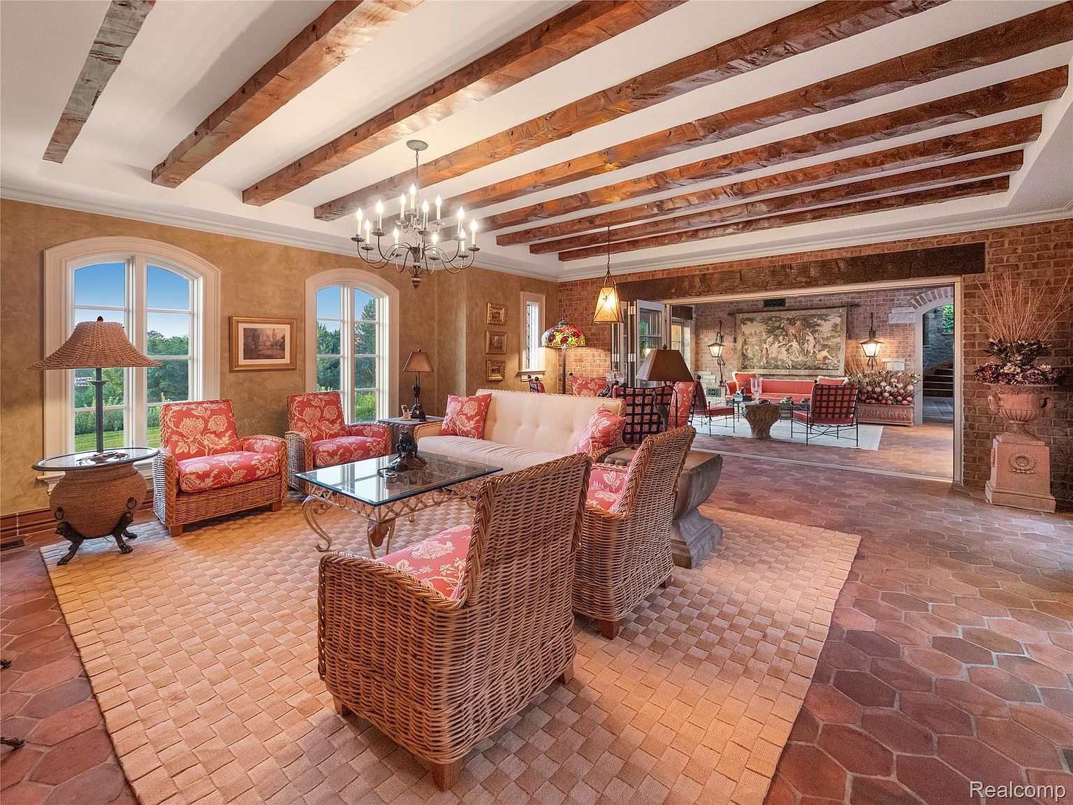 This is an interior shot of a luxurious living room featuring exposed wooden beams, a brick accent wall, and a mix of wicker and upholstered furniture. The room is well-lit with natural light from the windows and a chandelier, creating a warm and inviting atmosphere. The open layout leads to another seating area, enhancing the sense of spaciousness.