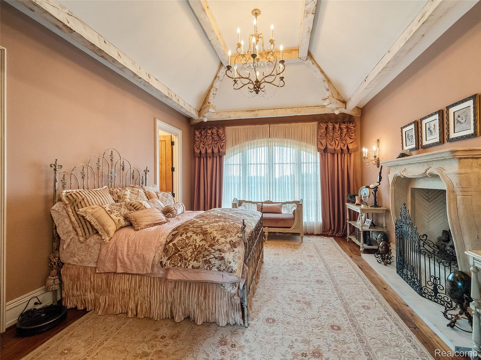 This is a luxurious primary bedroom featuring a wrought iron bed frame with ornate bedding, a stone fireplace with decorative ironwork, and a large arched window with elegant draperies. The room is decorated in a warm, romantic color palette with a chandelier providing soft lighting, creating an opulent and inviting atmosphere. The perspective is from the doorway, showcasing the depth and grandeur of the room.