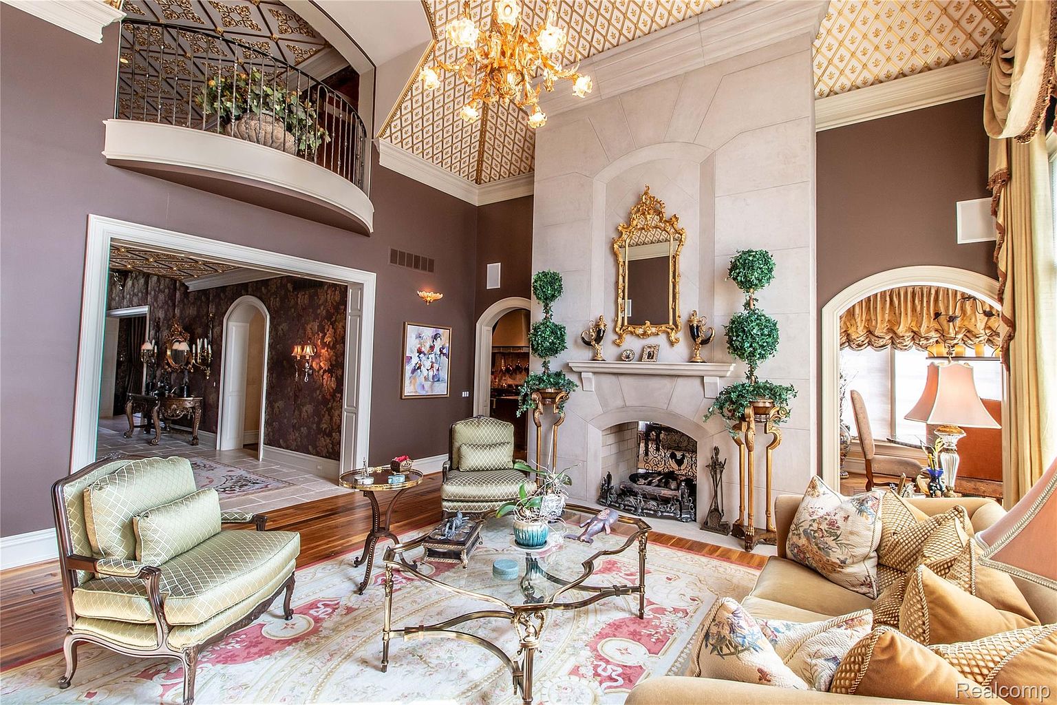 This is an interior shot of a luxurious living room featuring a high ceiling with intricate gold detailing, a grand stone fireplace with a gold-framed mirror, and elegant furnishings. The room is decorated in rich brown and gold tones, creating a warm and opulent atmosphere. The perspective is from the entrance of the room, showcasing the depth and grandeur of the space.