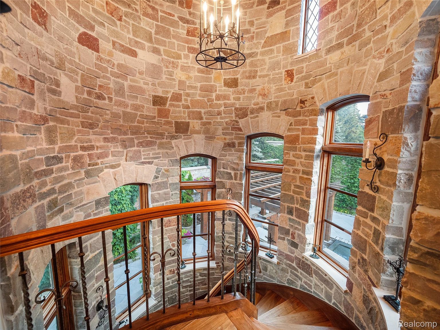 This image showcases a stunning spiral staircase within a stone tower. The staircase features wooden steps and an ornate metal railing, illuminated by a chandelier hanging from the ceiling. Several arched windows provide natural light and views of the surrounding landscape, creating a unique and elegant architectural feature.