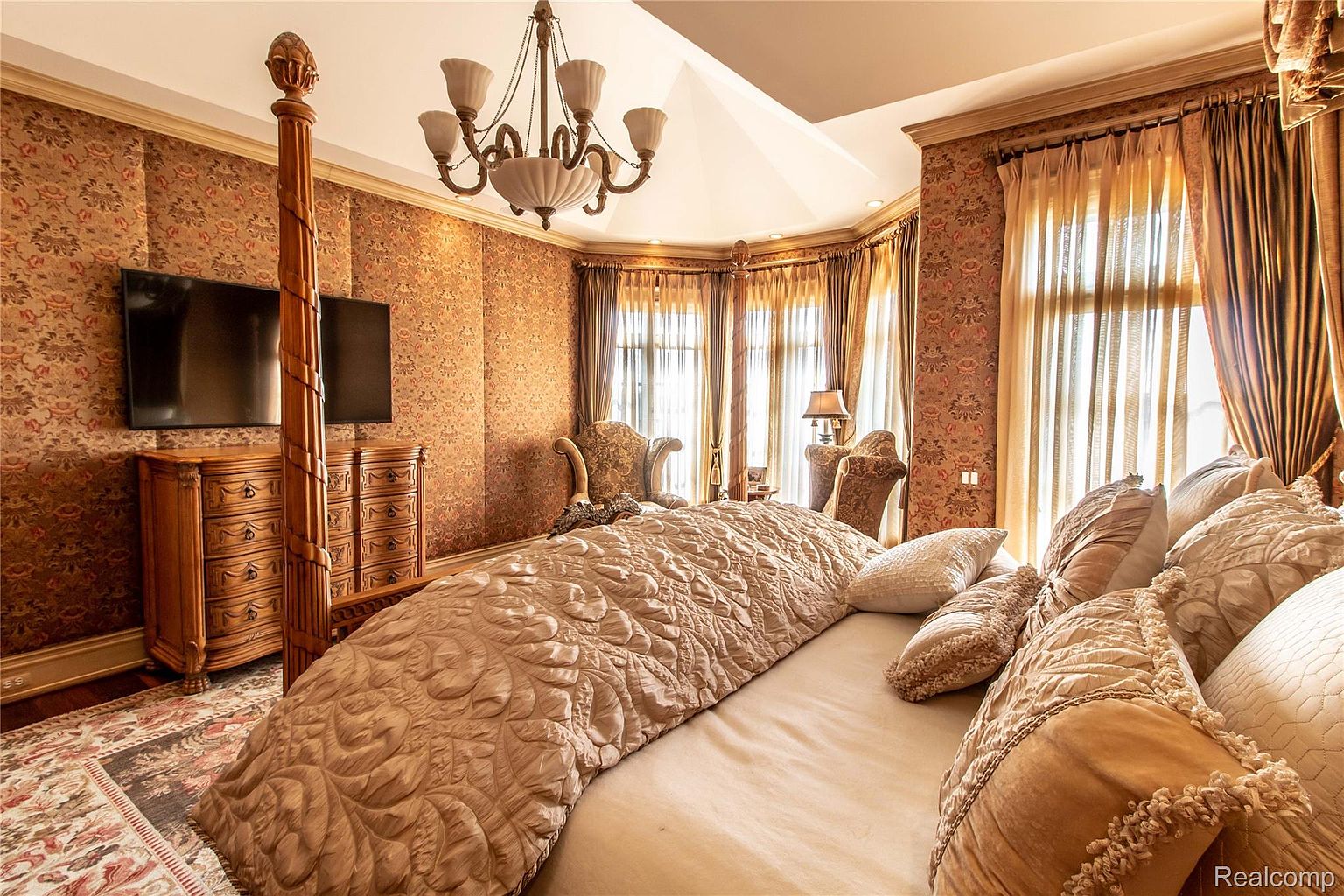 This is a luxurious primary bedroom featuring a four-poster bed with ornate detailing and a plush, patterned comforter. The room is decorated with rich, floral wallpaper and heavy drapes, creating a warm and opulent atmosphere. A chandelier hangs from the ceiling, and a television is mounted on the wall above a wooden dresser, adding modern convenience to the classic design.
