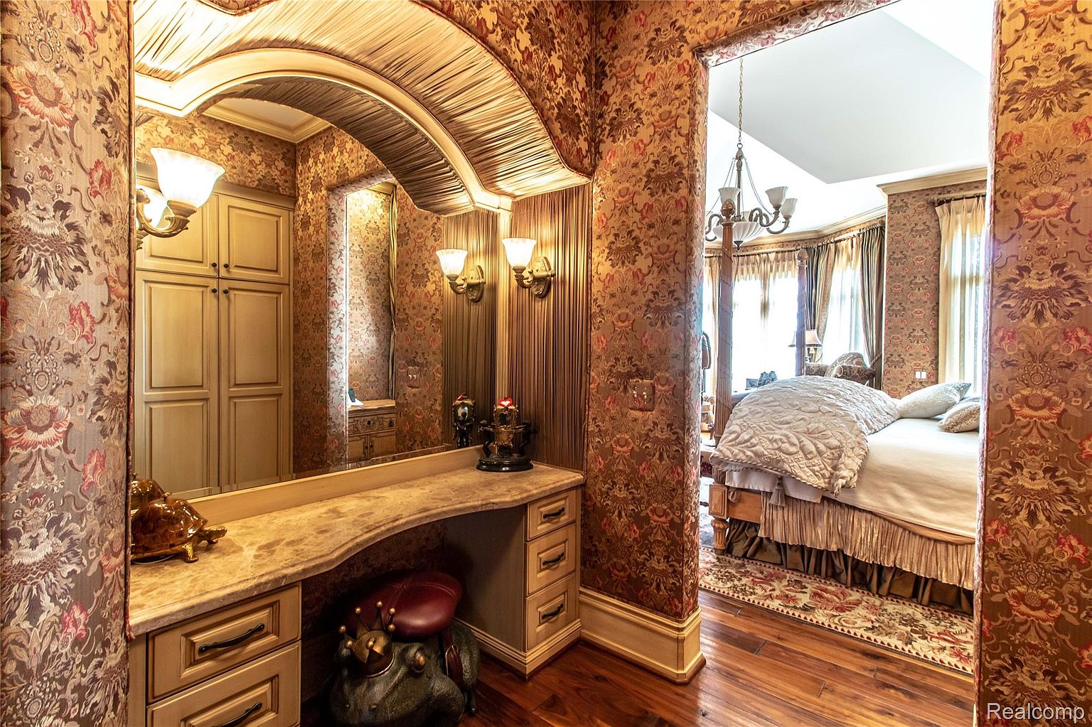 This image showcases a luxurious primary bedroom suite, featuring an ornate vanity area with a large mirror and custom cabinetry. The bedroom itself is visible through an archway, revealing a large bed with plush bedding, elegant drapery, and a chandelier. The room is decorated with rich, patterned wallpaper and hardwood flooring, creating a sophisticated and opulent atmosphere.
