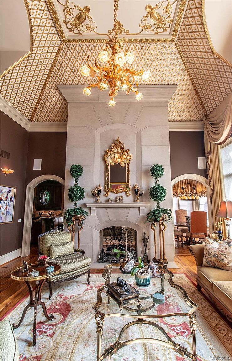 This is an opulent living room featuring a high, intricately patterned gold and white ceiling and a large stone fireplace as a focal point. The room is furnished with elegant, traditional-style furniture, including a patterned armchair, a glass-topped coffee table with gold accents, and a plush sofa. The color palette is rich and warm, creating a luxurious and inviting atmosphere.