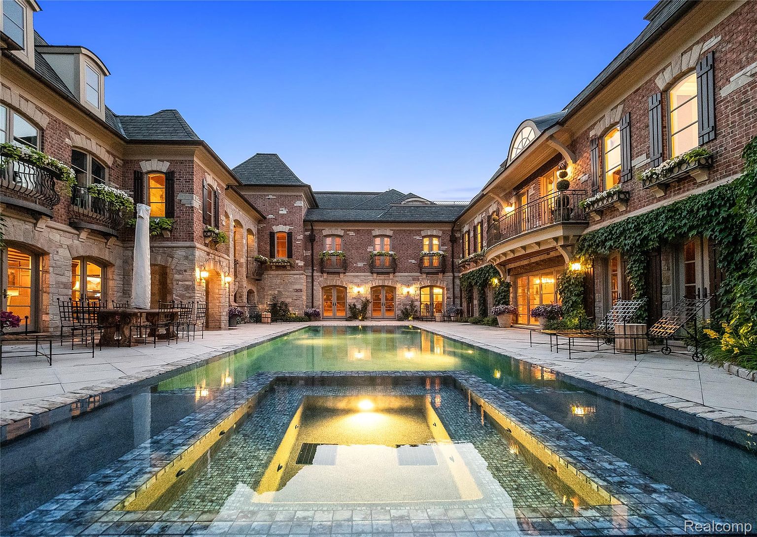 This image showcases a luxurious outdoor pool and spa area nestled within a courtyard of a grand estate. The pool features a unique design with a built-in spa, surrounded by elegant brick and stone architecture. The scene is bathed in soft evening light, creating a serene and inviting atmosphere, perfect for relaxation and entertainment.
