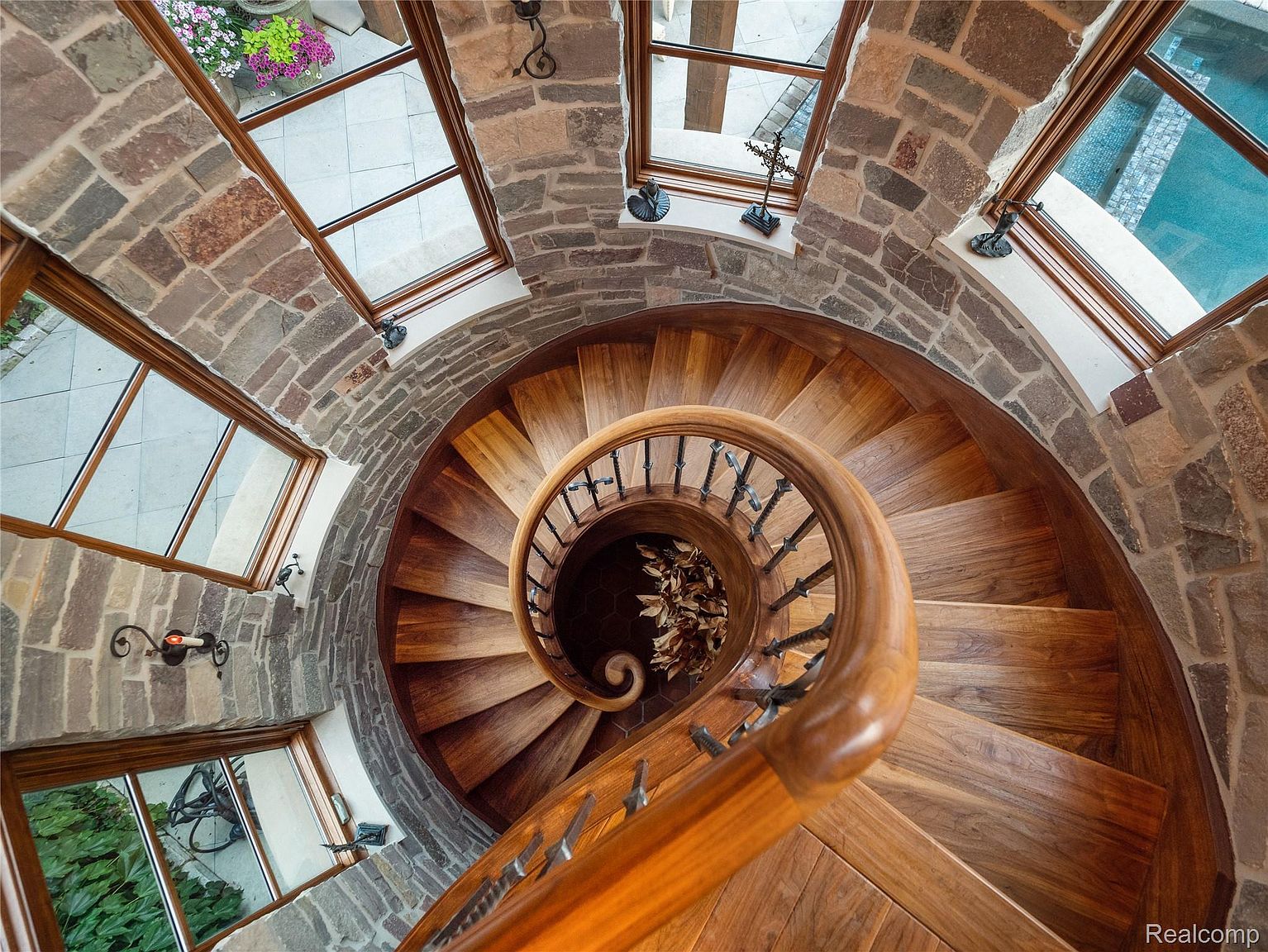 This image showcases a stunning spiral staircase constructed of wood, winding upwards within a stone-walled tower. Natural light streams in through multiple windows, highlighting the craftsmanship of the staircase and the rustic charm of the stone walls. The view is from above, looking down into the center of the spiral.