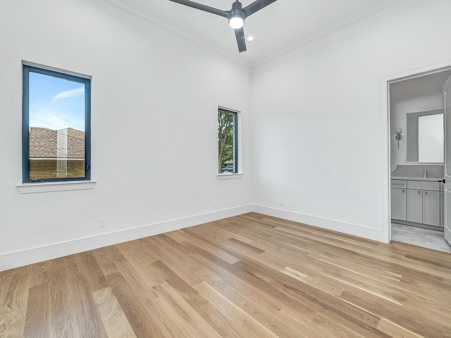 This is a bright and airy bedroom featuring hardwood floors, white walls, and two windows that provide natural light. The room has a minimalist aesthetic with clean lines and a neutral color palette. A glimpse into an adjacent bathroom reveals modern fixtures and finishes, enhancing the appeal of the space.
